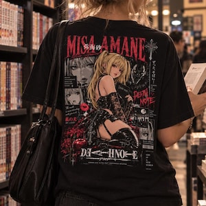 May include: A black t-shirt featuring a graphic print of the character Misa Amane from Death Note on the back. The design includes red and white text, a cross, a red apple, and multiple illustrations of the character in a dark aesthetic.
