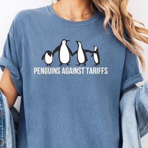 Könnte beinhalten: Ein blaues T-Shirt mit einer Grafik von vier Pinguinen, die sich an den Händen halten. Der Text "PENGUINS AGAINST TARIFFS" ist unter den Pinguinen aufgedruckt.