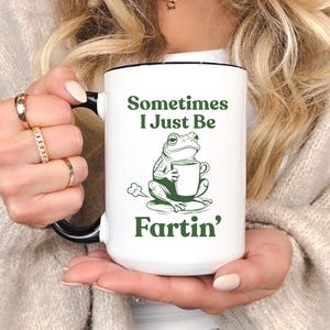 Pode incluir: Caneca de cerâmica branca com alça e borda pretas. A caneca apresenta o texto "Sometimes I Just Be Fartin'" com uma ilustração de um sapo verde segurando uma xícara de café. A caneca é segurada por uma pessoa.