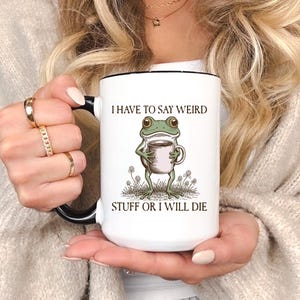 Op de afbeelding: Witte keramische mok met een zwarte handgreep en rand. De mok heeft een groene kikker die een koffiekopje vasthoudt, met de tekst "I HAVE TO SAY WEIRD STUFF OR I WILL DIE".