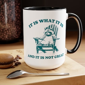 May include: White ceramic mug with a black rim and handle. The mug features a black and white illustration of a raccoon sitting in a green Adirondack chair. The text "It is what it is and it is not great" is printed in black on the mug.
