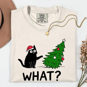 May include: Cream-coloured Comfort Colors XL t-shirt featuring a black cat wearing a Santa hat, gesturing towards a decorated Christmas tree. The text "WHAT?" is printed below the cat. A gold star pendant necklace is also present.