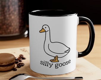 Könnte beinhalten: Weiße Keramiktasse mit schwarzem Rand und Henkel. Die Tasse zeigt eine schwarze Strichzeichnung einer Gans mit dem Text "silly goose" unterhalb der Gans.