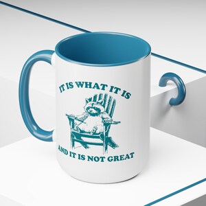 May include: White ceramic mug with a blue rim and handle. It features a raccoon sitting in a chair with the text "It is what it is and it is not great".