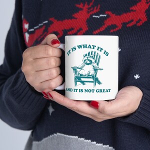 May include: White ceramic mug with a teal raccoon sitting in a teal Adirondack chair. The mug has the text "It is what it is and it is not great" printed on it.
