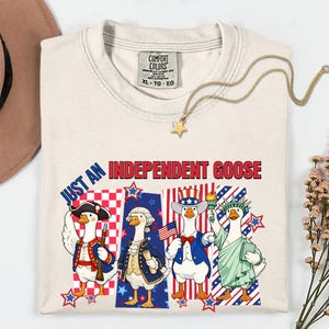 以下が含まれることがあります： 「JUST AN INDEPENDENT GOOSE」の文字と、愛国的な衣装を着た4羽のガチョウのグラフィックが描かれた、クリーム色のTシャツ。金の星のペンダントネックレスと茶色の帽子も見える。