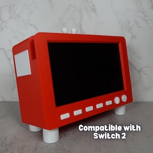 May include: A red rectangular device with a black screen, white buttons, and white cylindrical feet. The device has two white spheres on top and a white rectangular detail on the side. The text "Compatible with Switch 2" is visible on the bottom right.