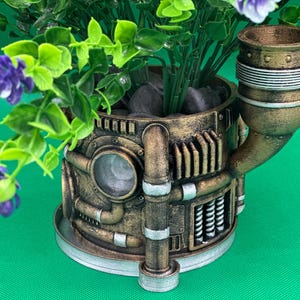 Może przedstawiać: Doniczka w stylu steampunk z brązowym, metalowym i srebrnym wykończeniem. Doniczka ma wylot w kształcie rury i okrągłą podstawę. Jest wypełniona zielonymi roślinami i fioletowymi kwiatami.