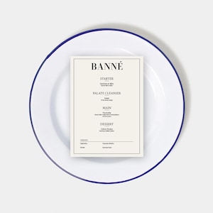 Puede incluir: Un plato de esmalte blanco con un borde azul, que presenta un menú impreso para un restaurante llamado "BANNÉ". El menú incluye entrante, limpiador de paladar, plato principal, postre y bebidas.