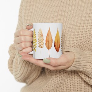 May include: White ceramic mug featuring a design of five stylised leaves in shades of yellow, orange, and brown. The leaves are arranged vertically down the mug.