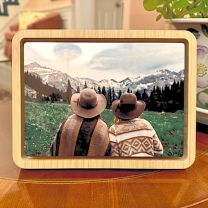 May include: Two people wearing hats stand back to back, looking out at a mountain range. The mountains are covered in snow and the sky is cloudy. The people are wearing brown and white patterned clothing.