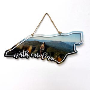 May include: A wooden cutout of the state of North Carolina with a photo of a mountain landscape and a person standing on a peak. The words "north carolina" are written in white cursive lettering on the cutout.