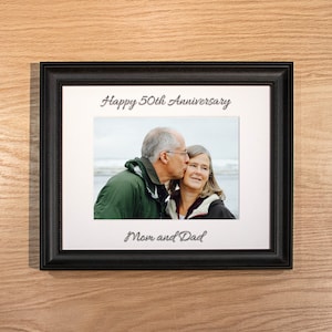 May include: A black picture frame with a white mat and a photo of an older couple. The photo shows the man kissing the woman on the cheek. The text "Happy 50th Anniversary" is printed above the photo and "Mom and Dad" is printed below the photo.