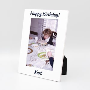 May include: White picture frame with a black stand. The frame has a photo of a child blowing out candles on a birthday cake. The text "Happy Birthday!" is printed on the top of the frame and the text "Kurt" is printed on the bottom of the frame.