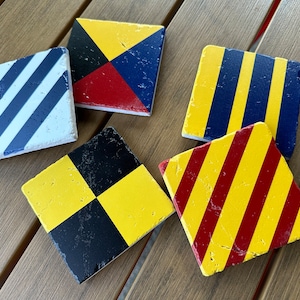 May include: Five square coasters with different nautical flag designs. The coasters are made of a light gray stone with a rough texture. The designs include yellow and red stripes, a checkered pattern of yellow and black squares, a blue and yellow striped pattern, and a red, yellow, and blue pattern.
