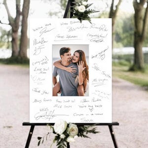 May include: A white rectangular guest book sign featuring a central color photograph of a person giving another person a piggyback ride. The white border is filled with various handwritten black ink signatures and messages of congratulations.