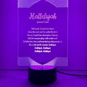 May include: A white acrylic light-up sign with a black base. The sign features the lyrics to the song "Hallelujah" by Leonard Cohen. The lyrics are: "Well people I've been here before I know this room and I've walked this floor You see I used to live alone before I knew ya And I've seen your flag on the marble arch But listen love, love is not some kind of victory march, no It's a cold and it's a broken Hallelujah Hallelujah, Hallelujah Hallelujah, Hallelujah"