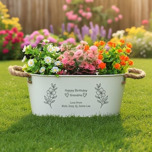 May include: A cream-coloured metal planter filled with colourful flowers, including white daisies, pink and orange blooms. The planter has rope handles and the text "Happy Birthday Grandma" with a floral design.
