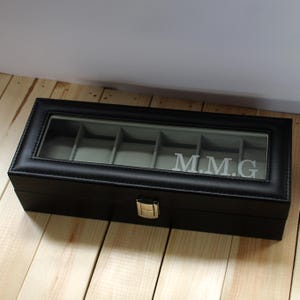 May include: Black leather watch box with a glass top and six compartments. The box has a silver latch and is engraved with the initials "M.M.G."