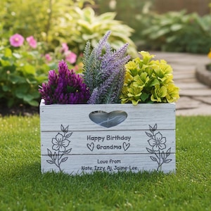 May include: A white wooden planter box filled with purple, lavender, and yellow flowers. The box has a heart-shaped cutout and the text "Happy Birthday Grandma" with a list of names below. Floral designs are on the sides.