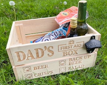 Op de afbeelding: Een houten kist met de tekst "Dad's First Aid Box" gegraveerd op de voorkant. De kist bevat een fles wijn, een fles sterke drank, een zak chips en een doos chocoladekoekjes. De kist heeft ook een flessenopener aan de zijkant.
