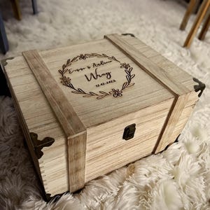 May include: A wooden keepsake box with a hinged lid and brass accents. The lid is engraved with a floral wreath design and the text "Emma & Andrew Wray 12.02.2023".