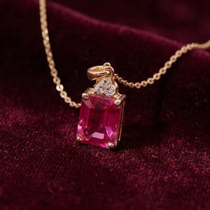 May include: A gold necklace featuring a rectangular, emerald-cut, vibrant pink gemstone pendant. The pendant is accented with three small, clear, round-cut stones at the top, set in a gold frame, and hangs from a delicate gold chain.