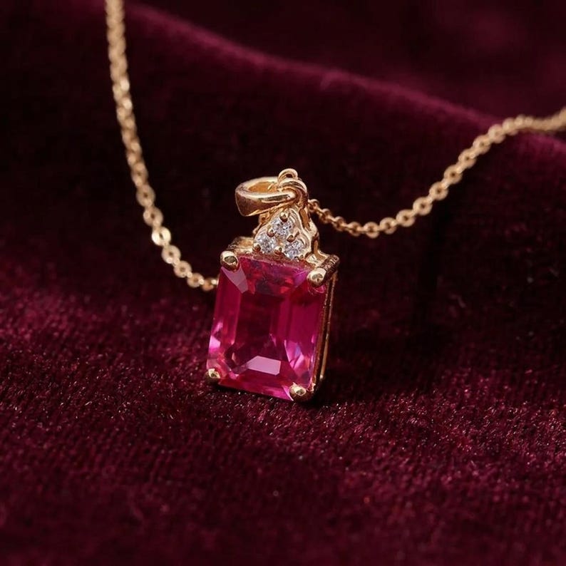 May include: A gold necklace featuring a rectangular, emerald-cut, vibrant pink gemstone pendant. The pendant is accented with three small, clear, round-cut stones at the top, set in a gold frame, and hangs from a delicate gold chain.