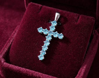 May include: A silver-toned cross pendant featuring multiple round, light blue faceted stones set in a vertical and horizontal arrangement. The pendant includes a small silver bail at the top for attachment, showcasing a bright, clear blue color.