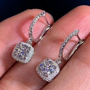 May include: A pair of silver drop earrings. Each earring has a square, clear gemstone surrounded by smaller, clear stones. The earrings have a hinged closure. The image shows the earrings against a blurred background.