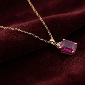 May include: A gold chain necklace featuring a rectangular emerald-cut magenta gemstone pendant. The pendant is accented with three small clear stones at the top, set in a gold frame that connects to a delicate gold link chain.