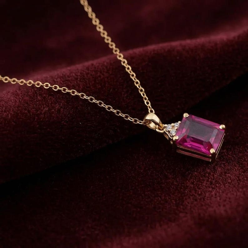 May include: A gold chain necklace featuring a rectangular emerald-cut magenta gemstone pendant. The pendant is accented with three small clear stones at the top, set in a gold frame that connects to a delicate gold link chain.