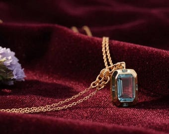 May include: A gold-toned necklace featuring a rectangular pendant with a large, light blue, emerald-cut gemstone at its center. The pendant is framed by a polished gold-colored metal setting and hangs from a delicate, matching gold-toned chain.
