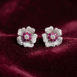 May include: A pair of flower-shaped stud earrings featuring a central clear round stone surrounded by a ring of deep pink stones, with petals entirely encrusted in small, shimmering white pavé stones set in a metallic frame.