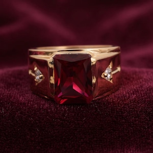 May include: A gold-toned ring featuring a large, rectangular, deep red gemstone set in the centre. The band is accented on each side by a small, triangular-shaped clear stone, providing a contrast to the prominent central red stone.