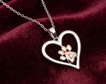 May include: A silver-toned heart-shaped pendant necklace featuring a rose gold-toned paw print cutout in the center. The paw print includes a small, clear, round gemstone set in the middle of the pad. The pendant hangs from a delicate silver chain.