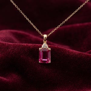 May include: A gold necklace featuring a rectangular, emerald-cut pink gemstone pendant. The pendant is accented with three small, clear, round-cut stones set in a triangular arrangement above the main pink stone, suspended from a delicate gold chain.
