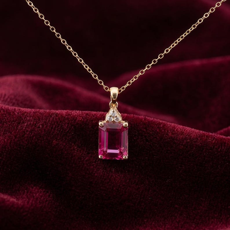 May include: A gold necklace featuring a rectangular, emerald-cut pink gemstone pendant. The pendant is accented with three small, clear, round-cut stones set in a triangular arrangement above the main pink stone, suspended from a delicate gold chain.