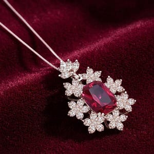 May include: A silver-toned necklace featuring a large, rectangular, deep red gemstone centerpiece. The pendant is surrounded by multiple small, clear, sparkling stones arranged in a floral pattern, attached to a thin, delicate silver-toned chain.