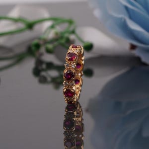 Puede incluir: Un anillo de oro con piedras preciosas rojas ovaladas alternas y piedras claras más pequeñas. El anillo se coloca sobre una superficie reflectante, con un fondo floral borroso. El diseño del anillo es simétrico y elegante.