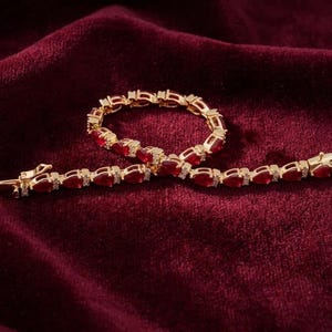 Könnte beinhalten: Ein goldfarbenes Armband mit einer Reihe von birnenförmigen roten Steinen, die sich mit kleinen klaren Akzentsteinen abwechseln. Das Armband ist mit einem sicheren Metallverschluss ausgestattet und zeigt ein durchgehendes Gliedermuster.