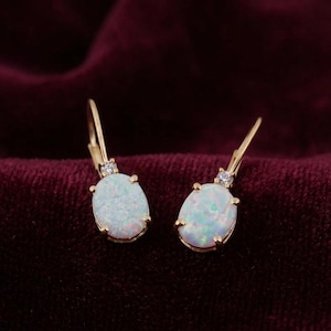 May include: A pair of gold leverback earrings featuring oval-shaped iridescent opals set in four-prong settings. Each earring is accented with a single small, clear round-cut gemstone positioned directly above the opal, adding a subtle sparkle.