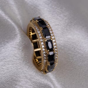 May include: A gold band ring featuring a row of black rectangular stones set in a pave setting. The ring is surrounded by a row of smaller clear stones.