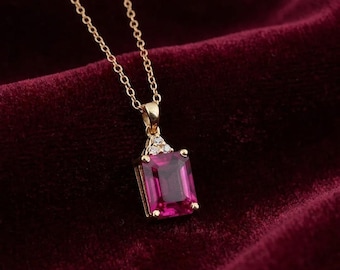 May include: A gold necklace featuring a rectangular emerald-cut magenta gemstone pendant. The pendant is accented with three small clear stones at the top where it connects to a delicate gold chain. The design is simple, elegant, and refined.