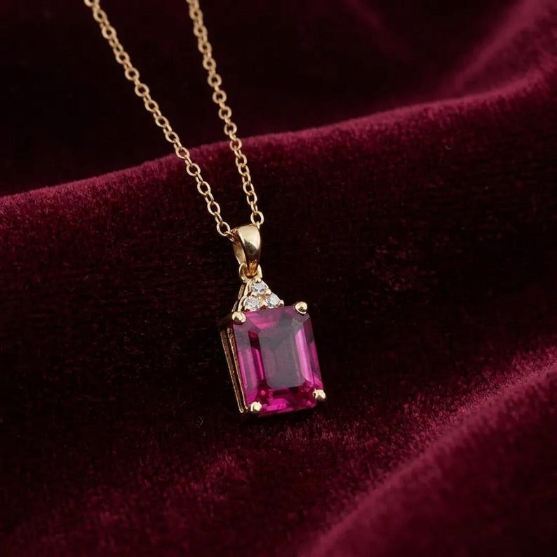 May include: A gold necklace featuring a rectangular emerald-cut magenta gemstone pendant. The pendant is accented with three small clear stones at the top where it connects to a delicate gold chain. The design is simple, elegant, and refined.