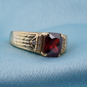 May include: A gold ring featuring a large, square-shaped, red gemstone surrounded by smaller, clear gemstones.