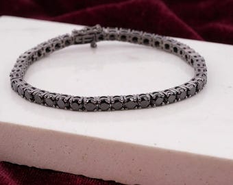 May include: A tennis bracelet featuring a continuous row of round-cut black stones set in a dark gunmetal-toned metal band. The bracelet includes a secure box clasp mechanism and is arranged in a circular shape on a flat white surface.