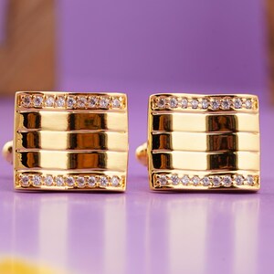 May include: A pair of gold cufflinks with a square design and a row of clear cubic zirconia stones around the edge.
