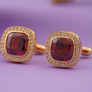May include: A pair of gold cufflinks featuring a square red gemstone set within a halo of smaller white gemstones.