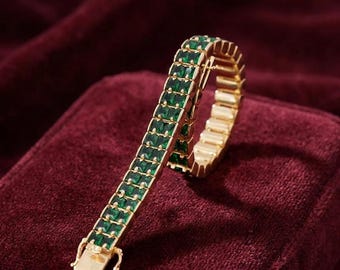 May include: A gold-toned tennis bracelet featuring a double row of square-cut emerald green stones. The bracelet has a flexible link construction and a rectangular metal clasp at the end, showcasing a uniform arrangement of vibrant green gems.